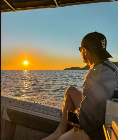 Person sitting on a boat looking at the sunset over water wearing 3D Embroided Black Cap.