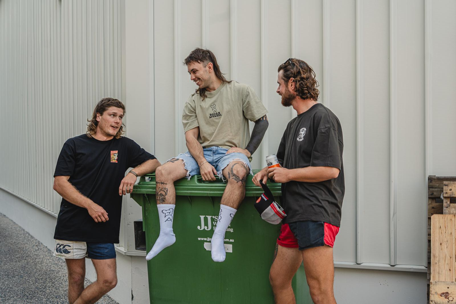 Three men sitting on a green dumpster with visible branding against a gray wall.
