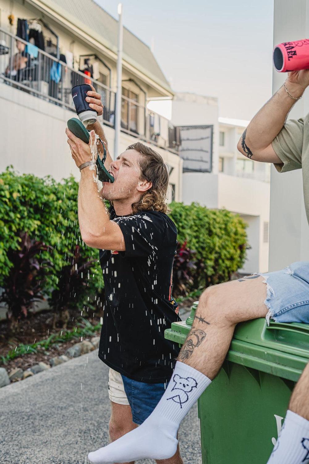 Two people sitting on a green trash bin, one doing a shoey. 